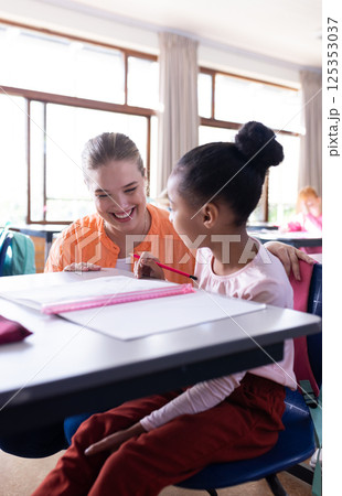 In school, female teacher smiling while helping young girl with writing assignment 125353037