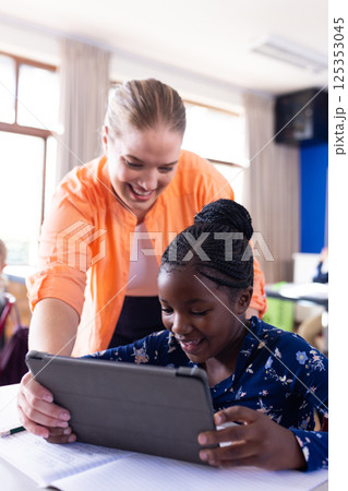 In school, female teacher helping african american girl using tablet in classroom, both smiling In school, female teacher helping african american girl using tablet in classroom, both smiling 125353045