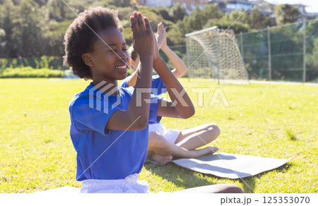 Practicing yoga on mats, diverse girls meditating outdoors at school field 125353070