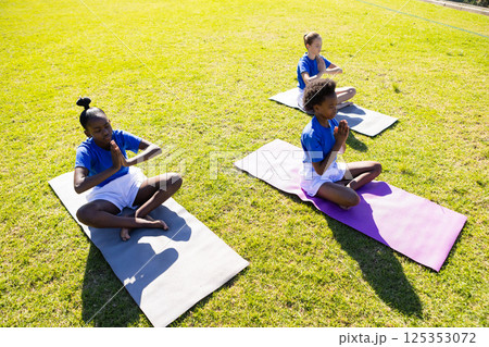 Practicing yoga on mats, three school diverse girls meditating outdoors on grass 125353072
