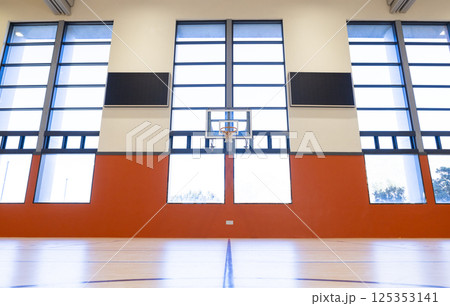 Basketball hoop in empty school gymnasium with large windows and high ceiling 125353141