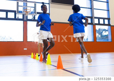 Running in school gym, diverse girls exercising around cones during physical education class 125353160