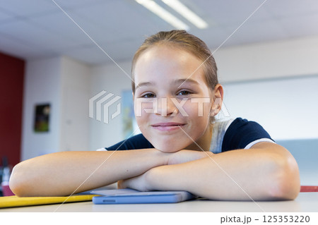 Smiling girl in school resting on desk with tablet, looking at camera 125353220