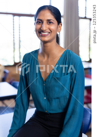 Smiling Indian female teacher sitting on desk in classroom, ready for school day 125353243