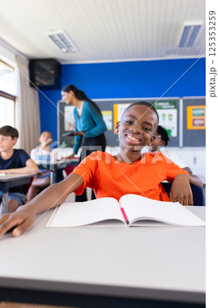 Smiling boy in classroom with open notebook, surrounded by classmates in school 125353259