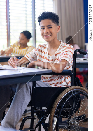 In school, boy in wheelchair writing in classroom, smiling at camera 125353297