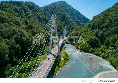 A suspension bridge stretches across a flowing river, surrounded by lush green mountains 125353582