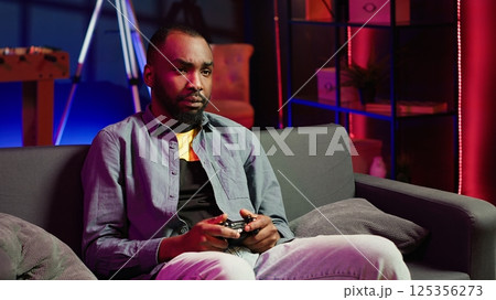 Focused gamer in RGB lit home gaming with gamepad, enjoying himself. Man sitting on sofa in neon illuminated living room, playing videogames on console using controller, camera A Focused gamer in RGB lit home gaming with gamepad, enjoying himself. Man sitting on sofa in neon illuminated living room, playing videogames on console using controller, camera A 125356273