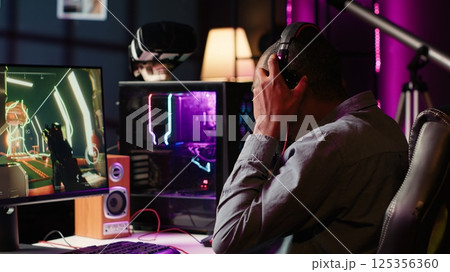 Smiling gamer with headphones on relaxing in gaming room, playing videogames on powerful computer, chatting with friends. Cheerful esports player performing in gaming tournament, camera A 125356360