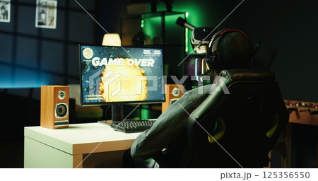 Heartbroken gamer dejected after losing in online multiplayer videogame, being defeated by rivals. Man putting hands on head in frustration after seeing game over screen on gaming pc monitor, camera B 125356550