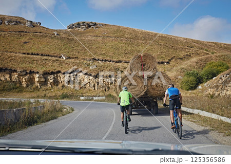 Two cyclists follow a truck loaded with hay bales along the road Two cyclists follow a truck loaded with hay bales along the road 125356586