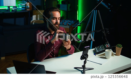 Influencer does technology review of wireless portable speaker for internet platforms channel, filming with mobile phone. Influencer showing music playing device, recording with smartphone, camera B 125356648