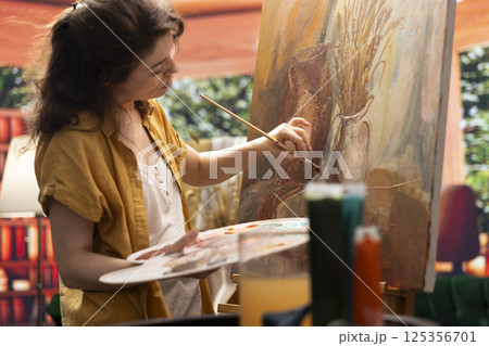 Female painter using watercolor paint and mixing colors on palette for her artistic creation in fine art workshop. Woman painting rustic elements on canvas, creative process in atelier. 125356701