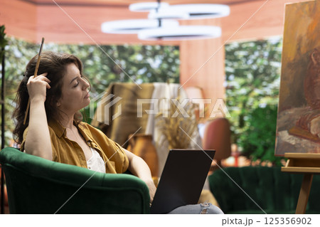 Female artist searching for a new painting inspiration on laptop, sitting in art and craft atelier and looking for creativity or imagination to create next masterpiece. Woman browses internet. Female artist searching for a new painting inspiration on laptop, sitting in art and craft atelier and looking for creativity or imagination to create next masterpiece. Woman browses internet. 125356902