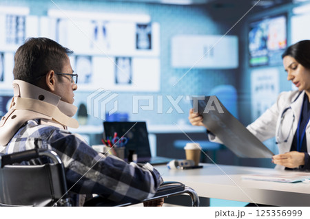 Indian medic reviewing x ray imaging test findings to patient with disability, recommending physiotherapy and professional wellness exercises. Female specialist assisting man with neck collar. 125356999