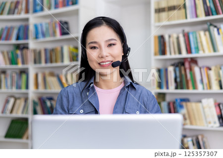 An attractive asian woman smiles while wearing a headset, working or studying remotely. The background shows bookshelves. 125357307