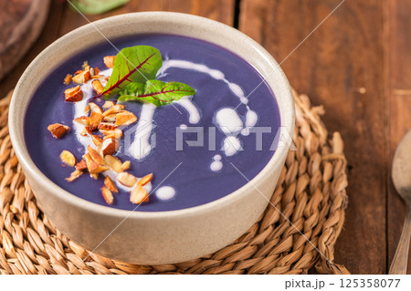 Purple soup garnished with chopped nuts, a drizzle of cream, and a green leaf, served in a beige bowl on a woven mat, placed on a wooden table 125358077