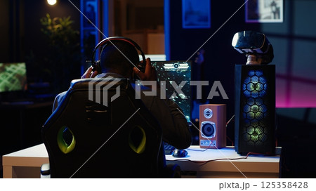 Man playing SF space videogame on PC screen in neon illuminated apartment living room. Gamer enjoying spaceship flying game on computer desktop display in RGB lit home late at night, camera A 125358428