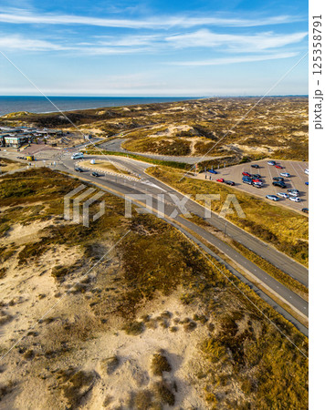 Aerial view of a coastal parking lot near sandy dunes with scattered cars, clear blue sky, and the sea in the distance. A road winds through the landscape leading to a beachside area. Aerial view of a coastal parking lot near sandy dunes with scattered cars, clear blue sky, and the sea in the distance. A road winds through the landscape leading to a beachside area. 125358791