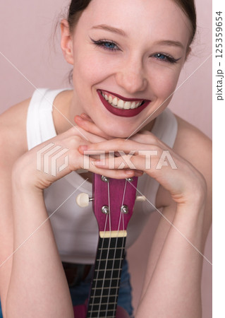 Portrait of young woman musician with ukulele. Beautiful 18 year old woman with big, toothy smile, positive attitude and joyful expression. She's head resting on her hands, cradling ukulele vertically Portrait of young woman musician with ukulele. Beautiful 18 year old woman with big, toothy smile, positive attitude and joyful expression. She's head resting on her hands, cradling ukulele vertically 125359654