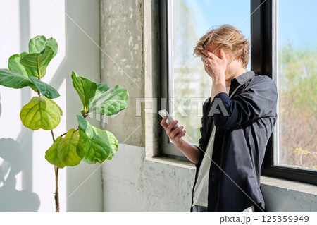 Upset sad teenager guy looking at smartphone screen, at home near window Upset sad teenager guy looking at smartphone screen, at home near window 125359949