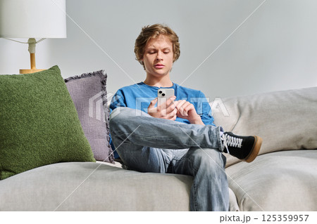 Handsome serious teenage guy using smartphone sitting on couch at home 125359957