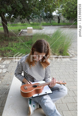 Girl Writes Notes On A Sheet Music To Play Them With The Ukulele 125360858