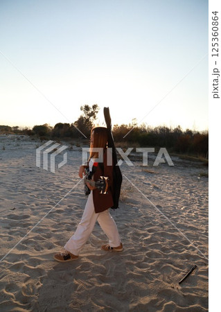 Girl Walks And Plays The Bass Instrument On The Beach Near The Ocean At Sunset 125360864