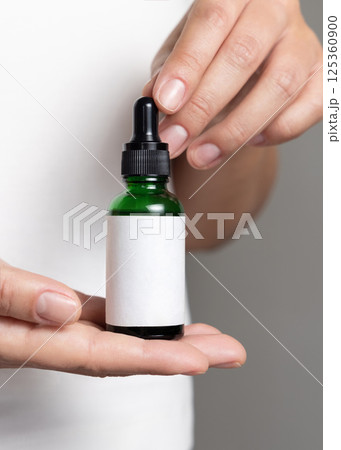 Female hands holding dropper bottle with a label against white shirt close up, cosmetic mockup 125360900