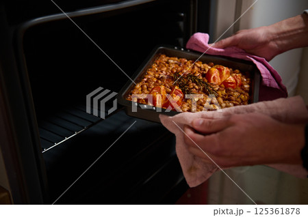 Carefully removing a baking tray of freshly cooked beans garnished with tomatoes and rosemary from the oven, evoking homely feelings of warmth and nourishment. Carefully removing a baking tray of freshly cooked beans garnished with tomatoes and rosemary from the oven, evoking homely feelings of warmth and nourishment. 125361878