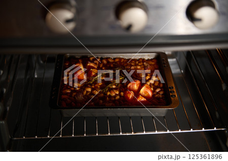 A baking tin filled with baked beans and tomatoes, flavored with rosemary, inside a preheated oven. The warm lighting enhances the inviting and appetizing nature of the dish during preparation. A baking tin filled with baked beans and tomatoes, flavored with rosemary, inside a preheated oven. The warm lighting enhances the inviting and appetizing nature of the dish during preparation. 125361896