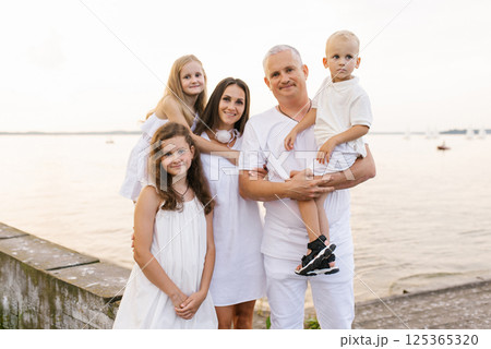 Family of five is posing for photo on the beach. Father is holding a baby, children are wearing white dresses 125365320