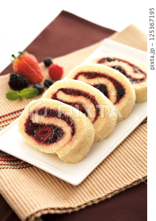 Close-up four slices of Swiss roll are on a white plate. The cake has a swirl of red filling. Berries and mint leaves are on the side. Close-up four slices of Swiss roll are on a white plate. The cake has a swirl of red filling. Berries and mint leaves are on the side. 125367195