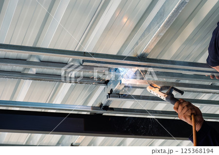 Metalworker welding steel frame of roof structure in industrial workshop Metalworker welding steel frame of roof structure in industrial workshop 125368194