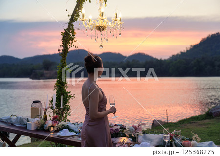 Beautiful asian woman standing holding a wine glass next to romantic dining table in the sunset Beautiful asian woman standing holding a wine glass next to romantic dining table in the sunset 125368275