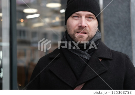 Handsome man wearing black coat and brown leather bag walking down on the city with mobile phone in hand. Stylish bearded middle-aged male using smartphone at urban street. Businessman going on job. 125368758