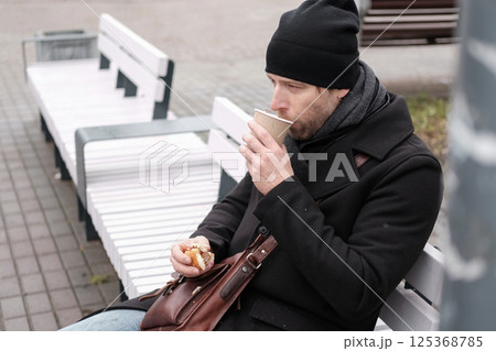 Pensive hipster drinking coffee sitting on a bench in the urban park. Handsome man wearing black coat eating street food, hot dog with paper cup in hand in the city. Stylish bearded middle-aged male. 125368785