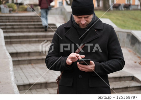 Handsome man wearing black coat and brown leather bag walking down on the city with mobile phone in hand. Stylish bearded middle-aged male using smartphone at urban street. Businessman going on job. 125368797