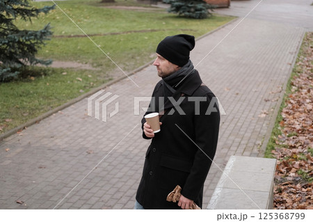Pensive hipster drinking coffee in the urban street. Handsome man wearing black coat, brown leather bag walking confidently down the city with paper cup in hand. Stylish bearded middle-aged male. 125368799