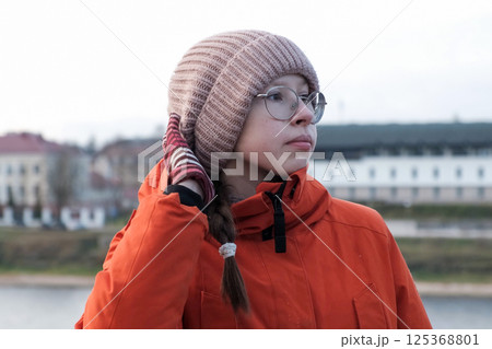 Teenager Girl Walking Alone on City Street. Portrait of 14 years old girl wearing eyeglasses, wool hat and winter jacket walks around the city. Urban Style. Rainy mood weather. Cold season. One person 125368801