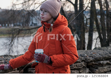 Teenager Girl Walking Alone on City Street. Portrait of 14 years old girl wearing eyeglasses, wool hat and winter jacket walks around the city. Urban Style. Rainy mood weather. Cold season. One person 125368804