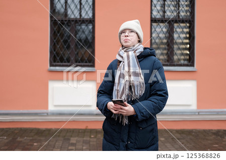 Teenager Girl Walking Alone on City Street with Smartphone. Portrait of 14 years old girl wearing eyeglasses, wool hat, winter jacket using mobile phone. Urban Style. Rainy mood weather. Cold season Teenager Girl Walking Alone on City Street with Smartphone. Portrait of 14 years old girl wearing eyeglasses, wool hat, winter jacket using mobile phone. Urban Style. Rainy mood weather. Cold season 125368826