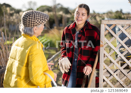 Two smiling casual women neighbors of different ages making arrangements during the vegetable garden season on sunny day of spring Two smiling casual women neighbors of different ages making arrangements during the vegetable garden season on sunny day of spring 125369049