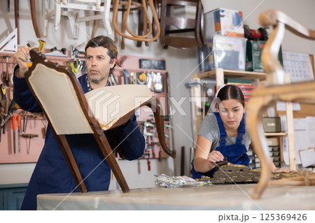 Middle-aged male restorer removes old cloth seat covering from antique wooden chair. Middle-aged male restorer removes old cloth seat covering from antique wooden chair. 125369426