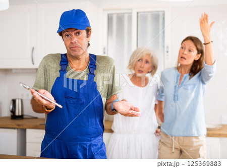 Inattentive plumber in uniform spreading his arms to the sides while being scolded by two women in the kitchen Inattentive plumber in uniform spreading his arms to the sides while being scolded by two women in the kitchen 125369883
