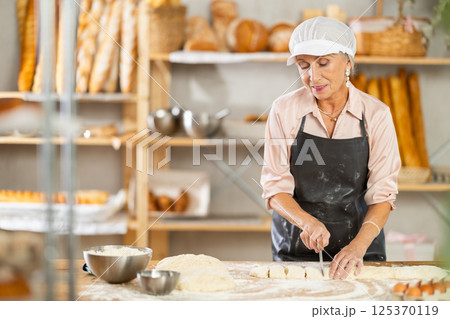 Senior woman works in bakery as baker, cuts dough into portions, form pieces of dough to create buns 125370119