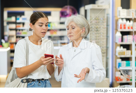 Senior female pharmacist communicates with girl client, looks at mobile phone screen 125370123