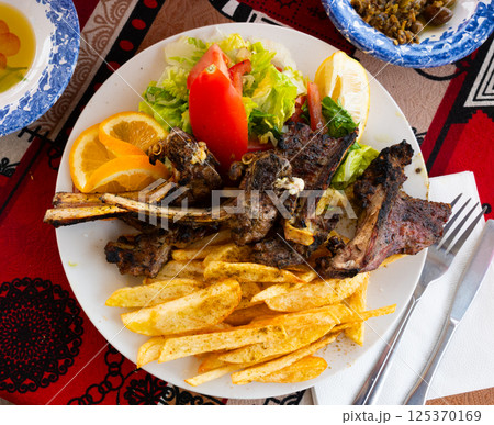 Delicious browned grilled lamb ribs served with crispy french fries and lettuce with tomato on plate, decorated with lemon. Delicious browned grilled lamb ribs served with crispy french fries and lettuce with tomato on plate, decorated with lemon. 125370169