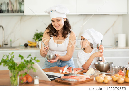 Happy mother and daughter wearing chef hats preparing salmon in the kitchen 125370276