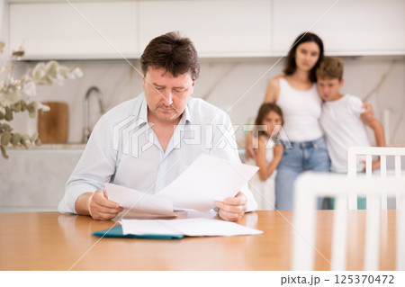 Upset father reads and looks through financial documents. Sad wife with children in background 125370472
